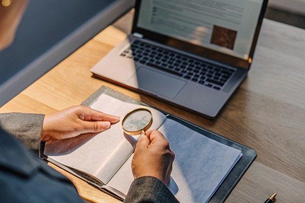 Person using magnifying glass on book near laptop (ID#2)